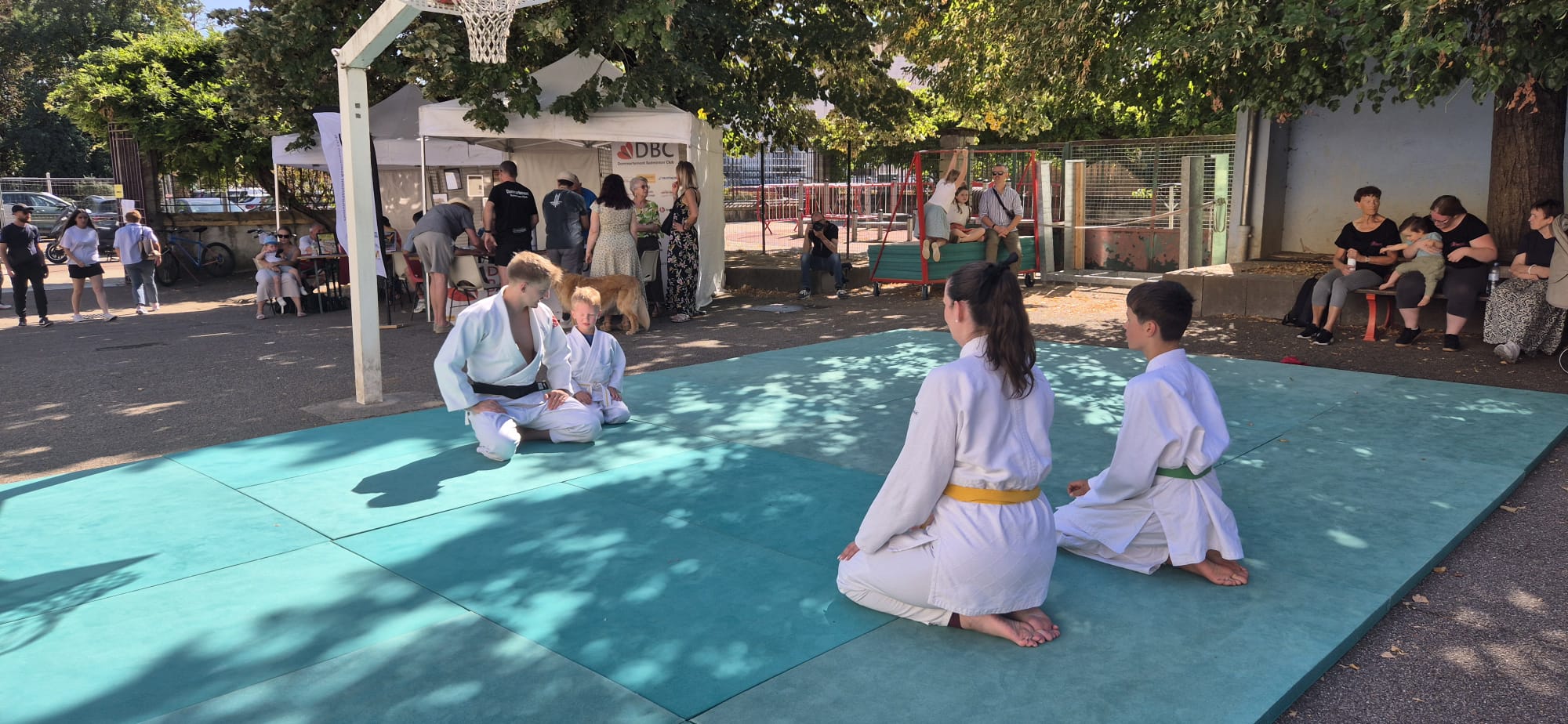 Le tatami vert du club installé dans la cour de l'école, quatre judokas assis en seiza, les stands du forum en arrière-plan